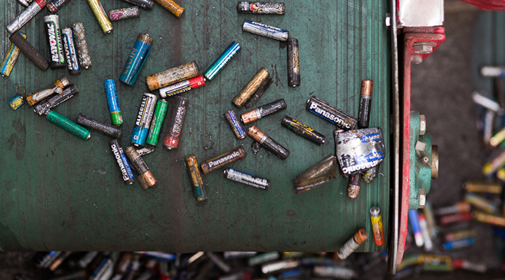 Foto, tirada de cima, de esteira verde de reciclagem com pilhas velhas e/ou sem uso. Em segundo aparecem algumas pilhas espalhadas no chão.