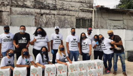 Pessoas enfileiradas lado a lado posam para foto; Caixas brancas com desenhos de árvores estão no chão, à frente delas,