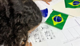 Foto de criança debruçada sobre desenho para colorir. Na mesa estão espalhados alguns papéis e cartazes em branco e pequenas bandeiras do Brasil.