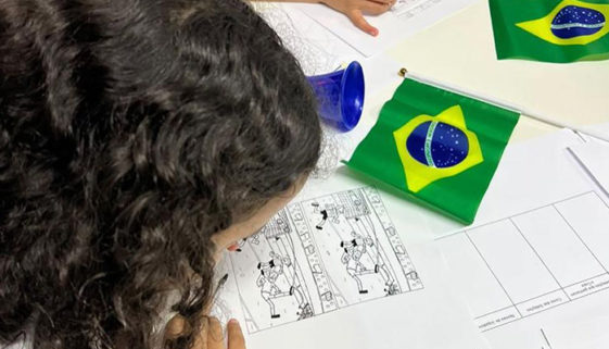 Foto de criança debruçada sobre desenho para colorir. Na mesa estão espalhados alguns papéis e cartazes em branco e pequenas bandeiras do Brasil.