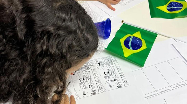 Foto de criança debruçada sobre desenho para colorir. Na mesa estão espalhados alguns papéis e cartazes em branco e pequenas bandeiras do Brasil.