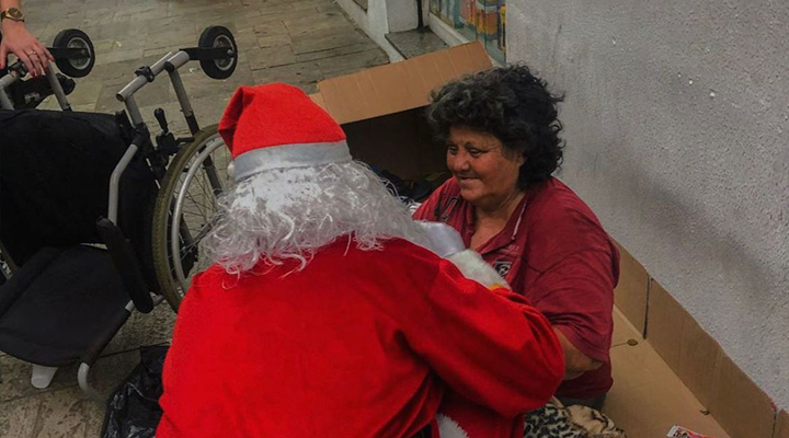 Foto de pessoa vestida de Papa Noel, agachada, interagindo com mulher em situação de rua, sentada em papelões.