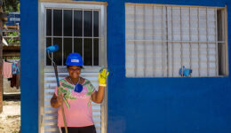 Foto de mulher parada à frente de porta de alumínio de casa pintada em azul. Ela veste um capacete, também de cor azul e posa sorridente para foto, segurando em sua mão direita um rolo de pintar. Com sua mão esquerda, vestida por uma luva amarela, ela acena.