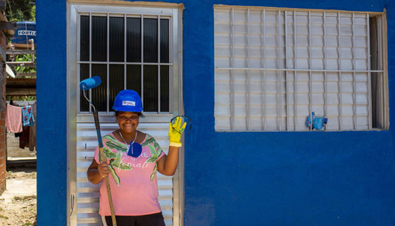 Foto de mulher parada à frente de porta de alumínio de casa pintada em azul. Ela veste um capacete, também de cor azul e posa sorridente para foto, segurando em sua mão direita um rolo de pintar. Com sua mão esquerda, vestida por uma luva amarela, ela acena.