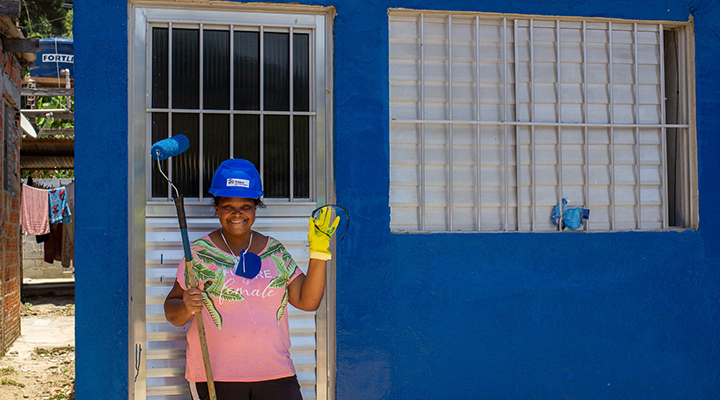 Foto de mulher parada à frente de porta de alumínio de casa pintada em azul. Ela veste um capacete, também de cor azul e posa sorridente para foto, segurando em sua mão direita um rolo de pintar. Com sua mão esquerda, vestida por uma luva amarela, ela acena.