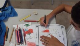 Foto de criança, vestida de uniforme, pintando desenho feito a mão. Sobre a mesa, estão outros desenhos e demais lápis de cor.