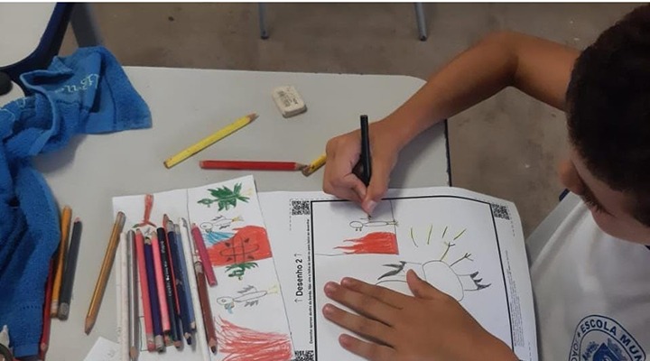 Foto de criança, vestida de uniforme, pintando desenho feito a mão. Sobre a mesa, estão outros desenhos e demais lápis de cor.