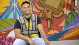 Foto de homem sorridente. Ele está sentado e veste um colete de trabalho. Ao fundo está uma parede com várias pinturas coloridas.