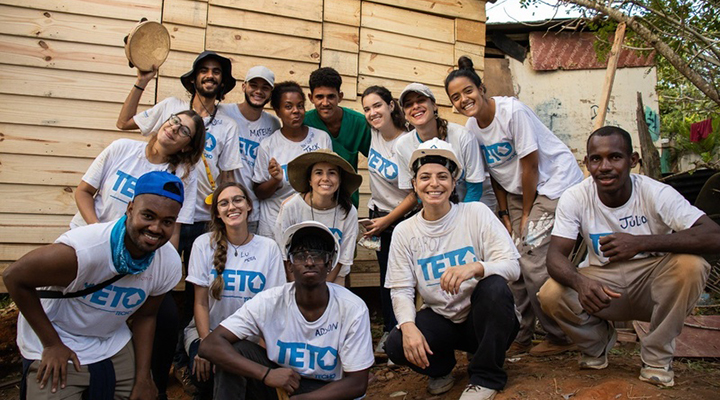 Foto de várias pessoas sorrindo e vestindo camisetas brancas com o logo da 'Teto' em azul.