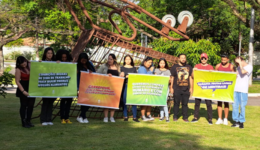 Foto de 12 jovens, lado a lado, segurando cartazes coloridos com frases de afirmação, em um gramado, à luz do dia.