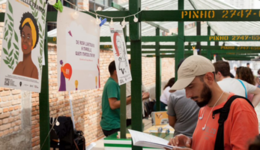 Foto de homem branco, usando boné bege e camiseta laranja folheando um livro; à frente dele estão algumas imagens penduradas em um varal. Está de dia.