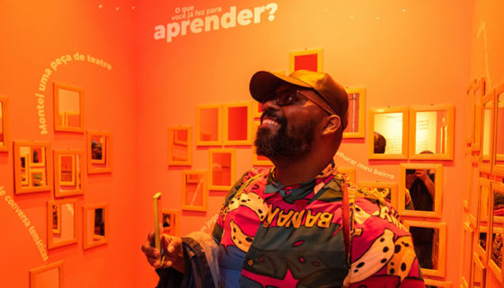 Foto de homem negro interagindo com sala de paredes laranjas. Ele está sorrindo enquanto segura o celular.