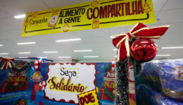 Foto de gôndola de supermercado, improvisada com papéis de temas natalinos, placas como o nome da campanha "Alimento a Gente Compartilha", "Seja Solidário", e "Doe"; e demais ornamentos temáticos.